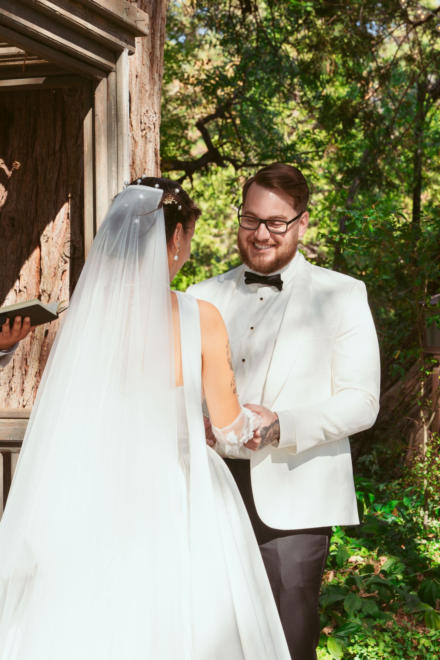 A couple shares an intimate first look moment under a rustic wooden pergola surrounded by greenery.