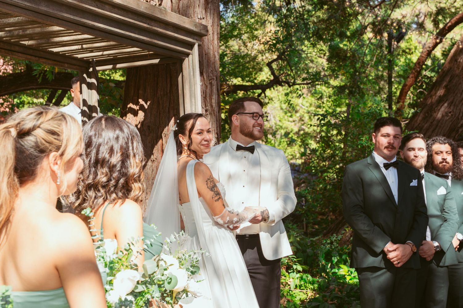 A wedding ceremony in progress under a wooden pergola surrounded by lush greenery.