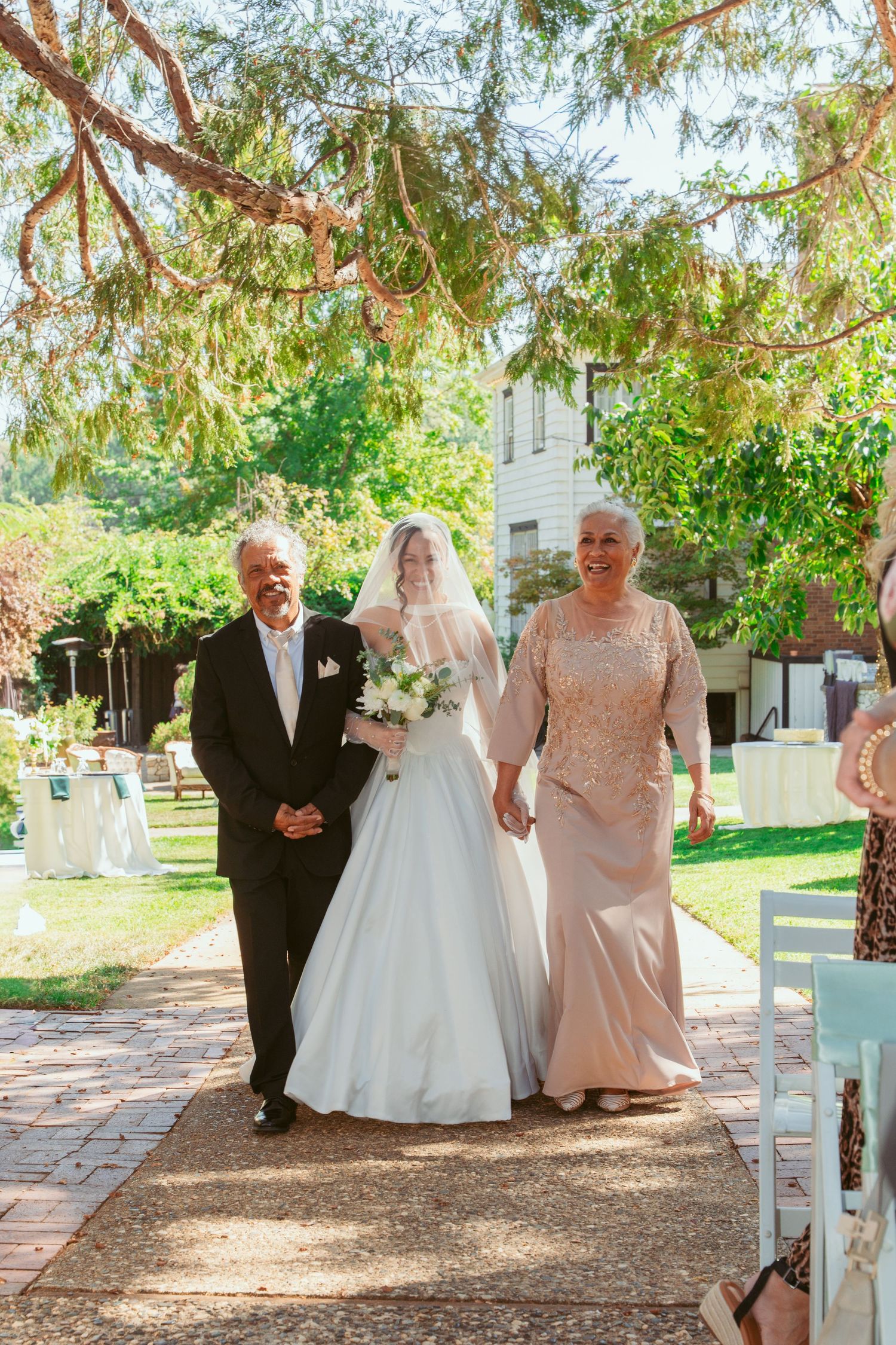 Wedding party walks along a tree-lined brick path at an outdoor ceremony venue.