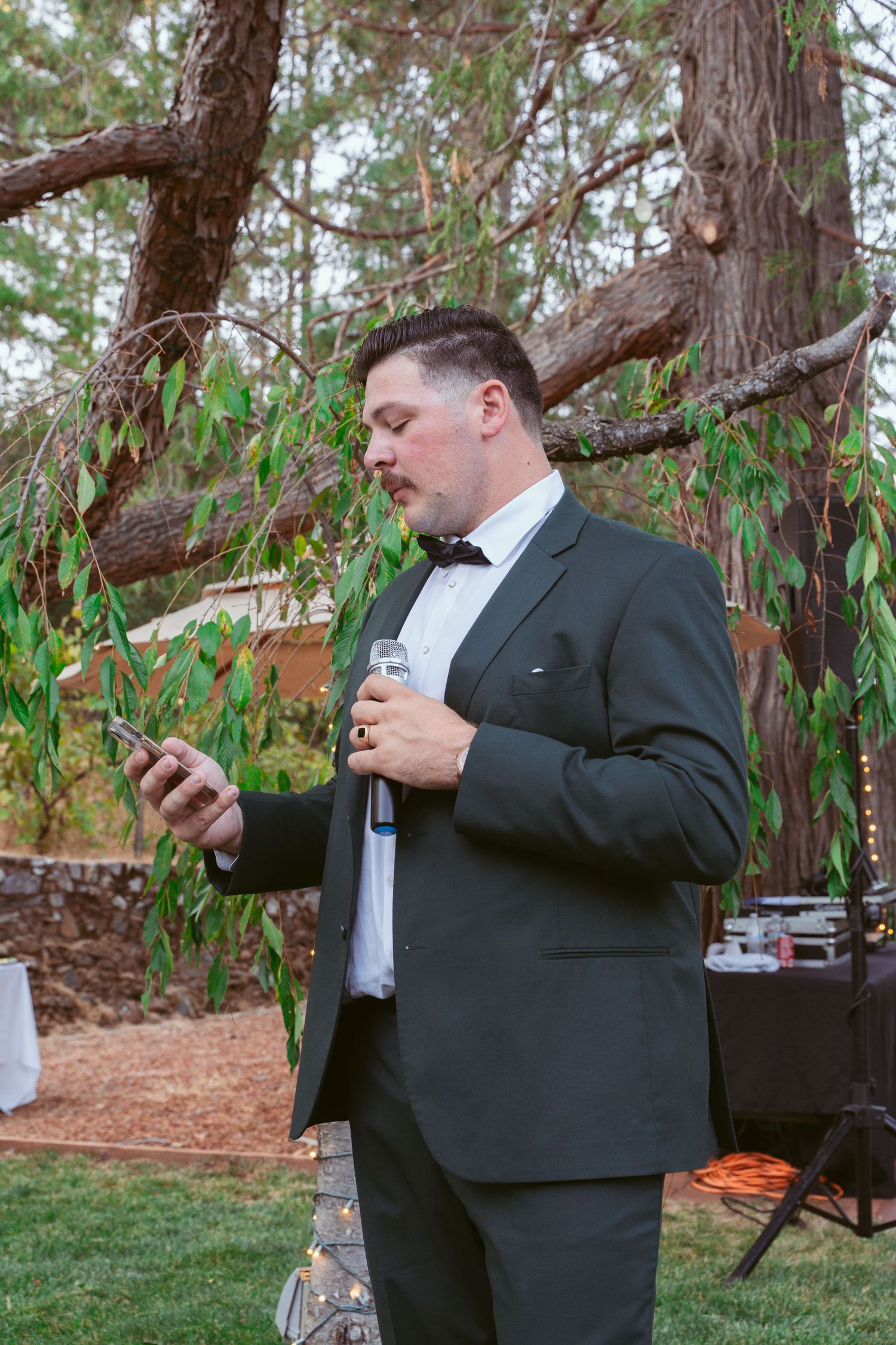 A person in a black suit checks their phone while standing in an outdoor wooded area.