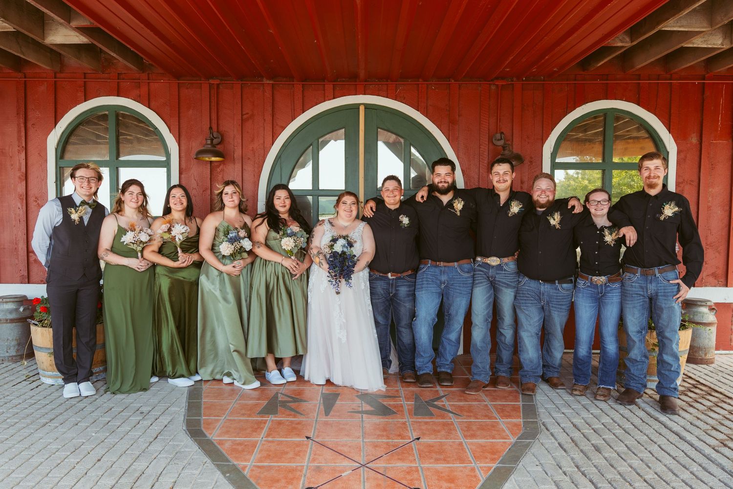 Large wedding party group photo in front of rustic red building with teal doors.