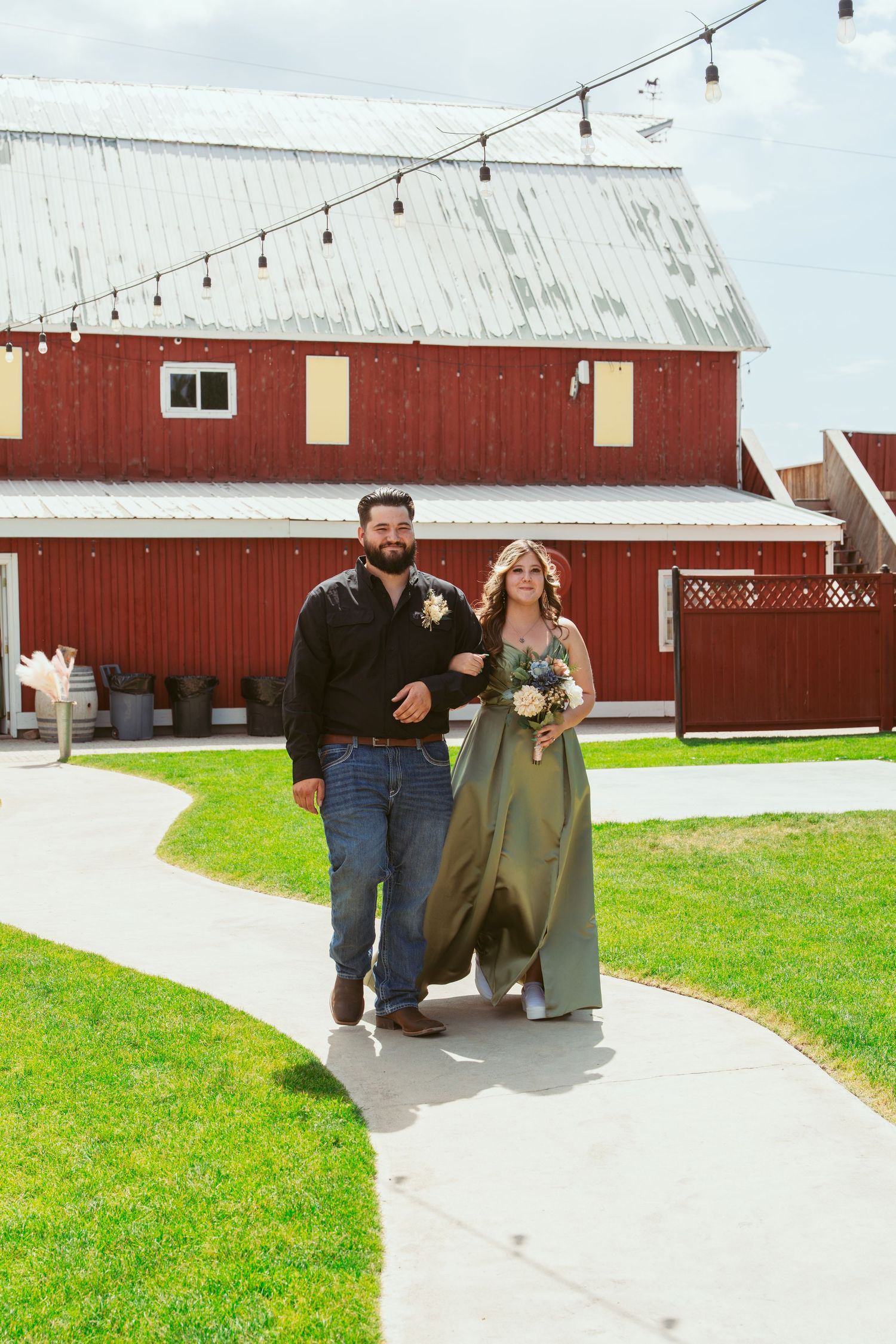 Couple walking along concrete path outside red barn venue with string lights overhead.
