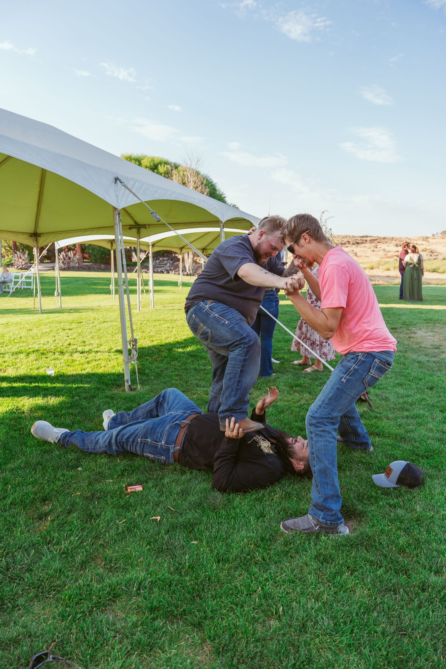 Playful group wrestling and roughhousing on grass near outdoor event tents.