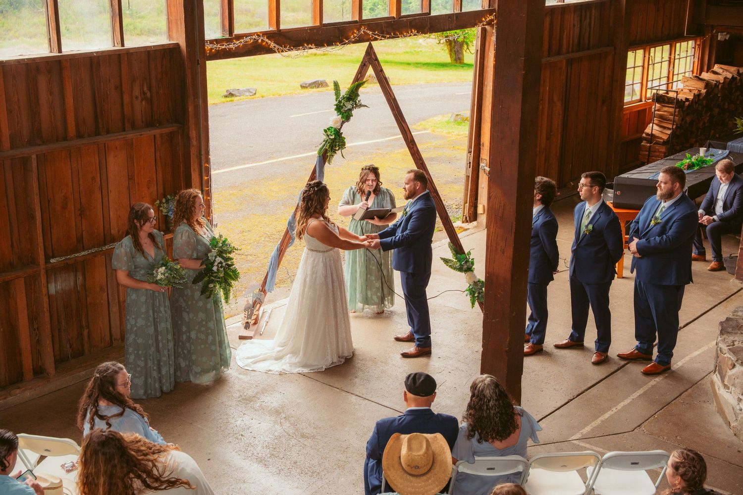 Wide angle view of rustic barn wedding ceremony with guests seated on wooden chairs.