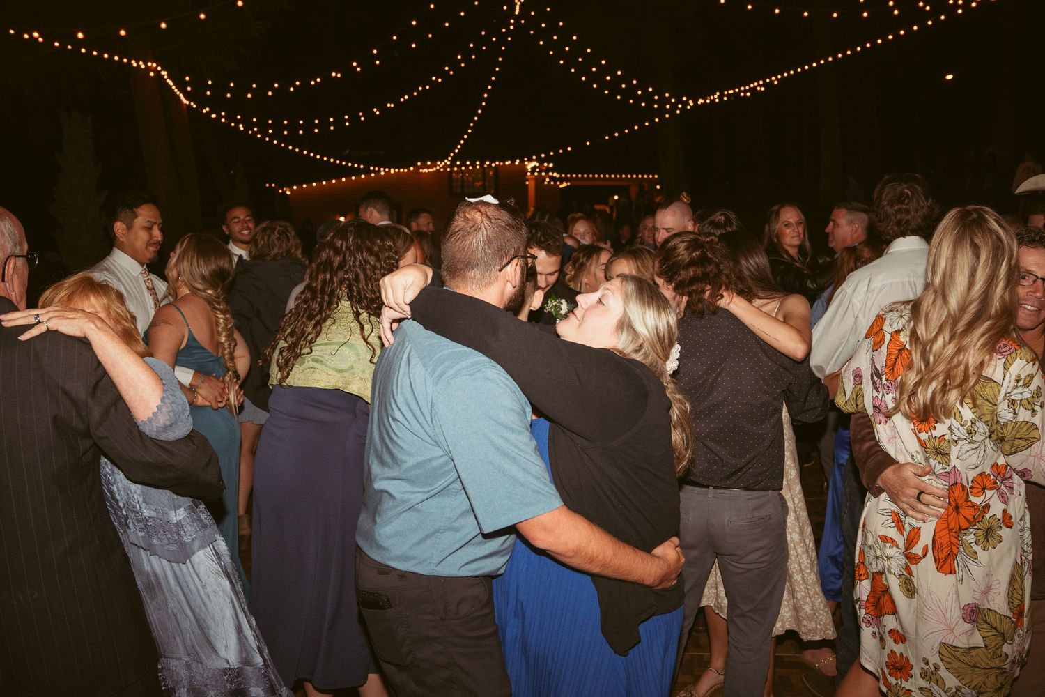 Wedding reception dance floor filled with guests celebrating under warm string lights.