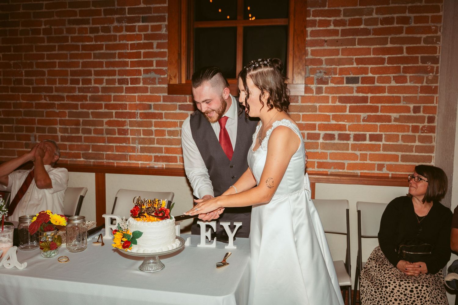 Wedding reception scene showing cake cutting with guests watching in a rustic brick venue.
