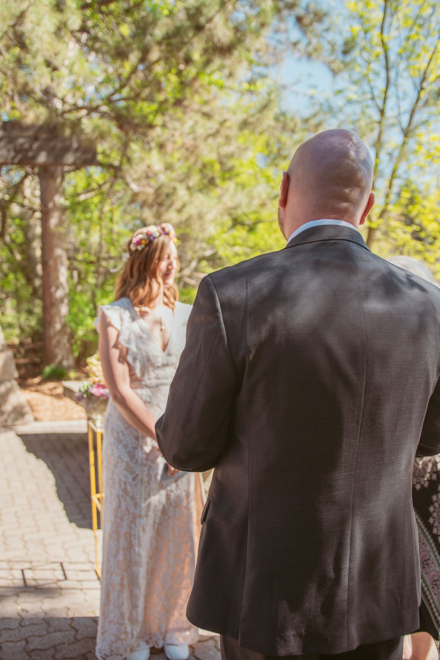 Wedding couple stands together on stone pathway surrounded by sunlit trees and greenery.