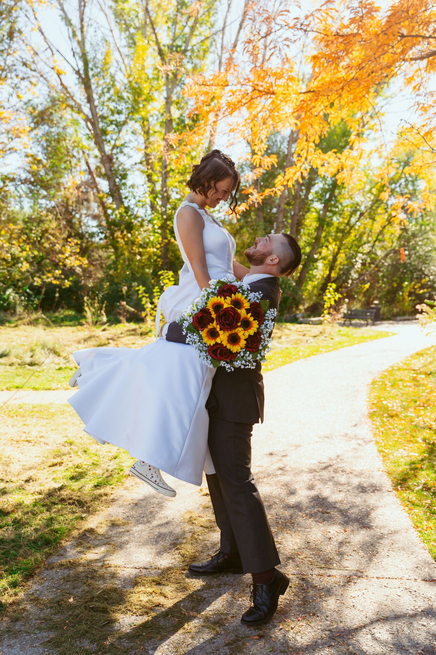 A couple shares a romantic moment with sunflowers on a path surrounded by vibrant fall colors.