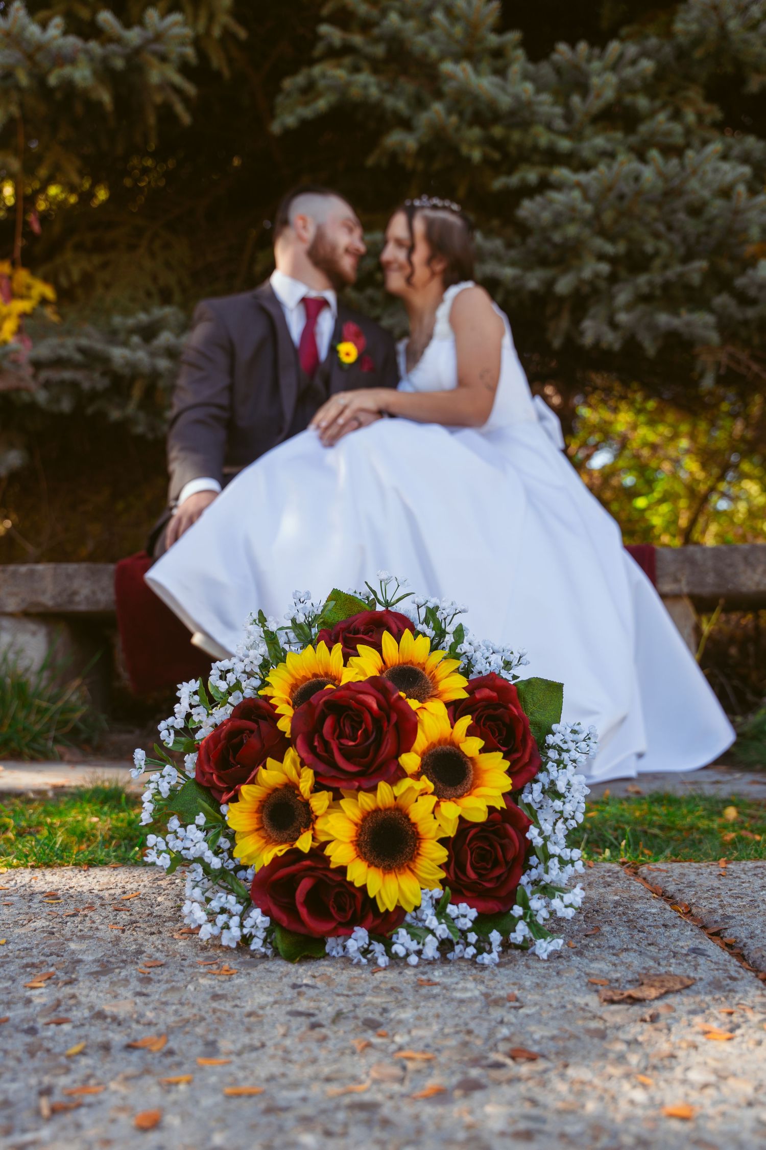 Wedding bouquet with sunflowers and burgundy roses rests on ground while couple poses in background.