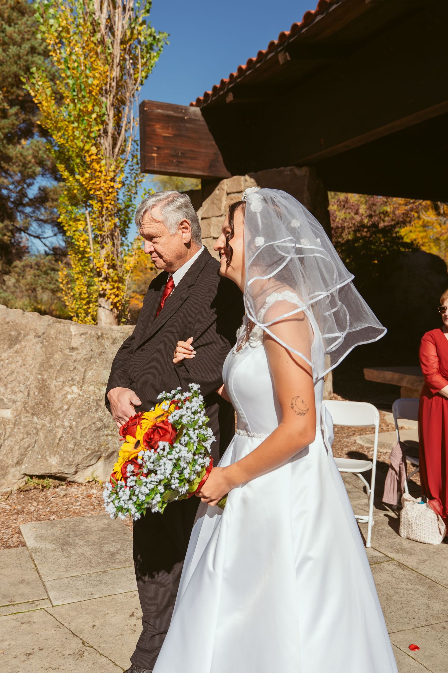 Bride in white dress and veil walks outdoors during autumn wedding ceremony.
