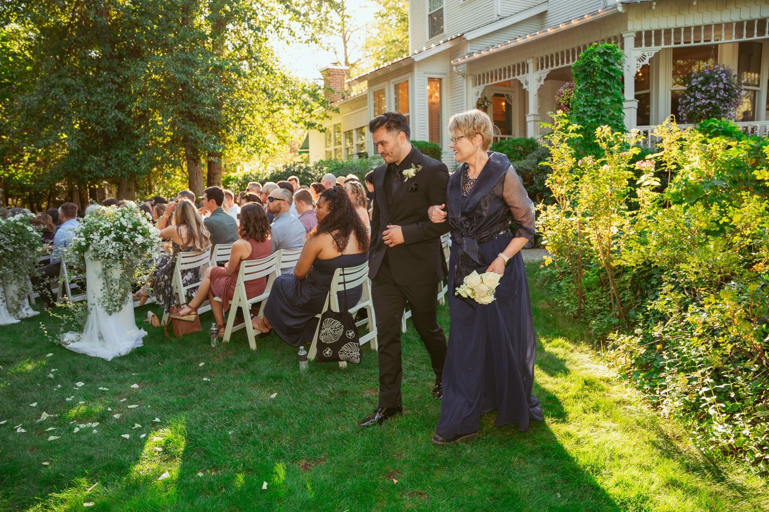Wedding guests seated on white chairs during an outdoor garden ceremony at sunset.