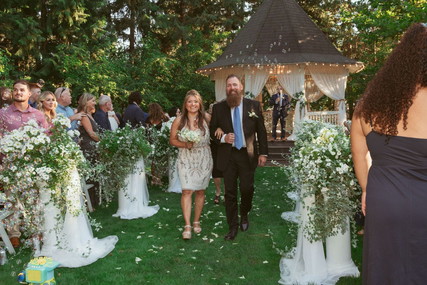 Couple walks down outdoor aisle decorated with white floral arrangements near garden gazebo.