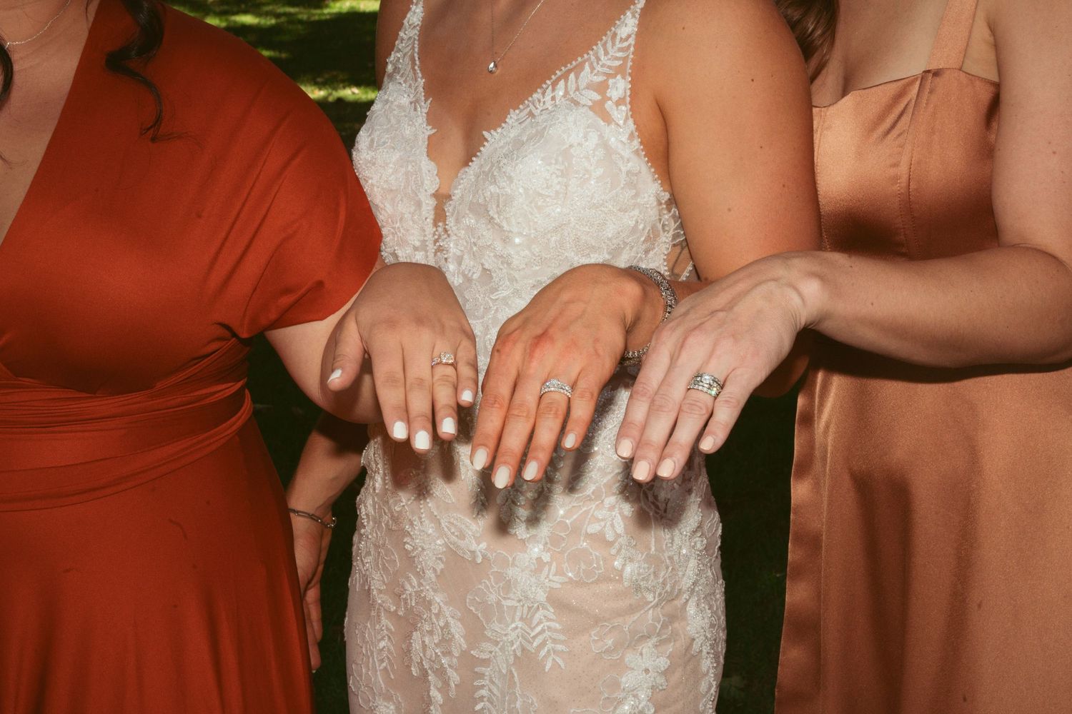 Close-up of hands showing wedding rings with a white lace dress and rust-colored bridesmaid dresses.