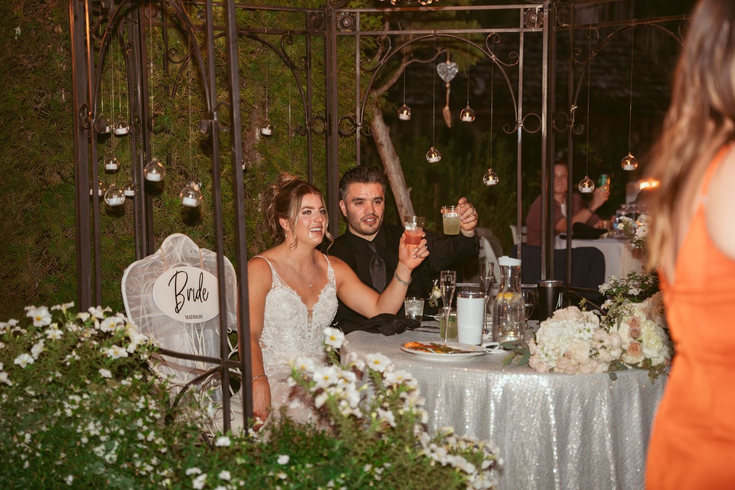 Outdoor wedding reception venue with hanging lights and white floral centerpieces on round tables.