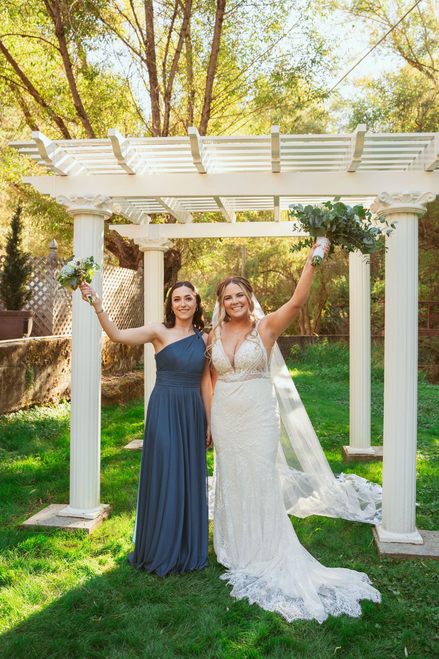 Two people in formal attire pose playfully in front of a white pergola surrounded by lush greenery.