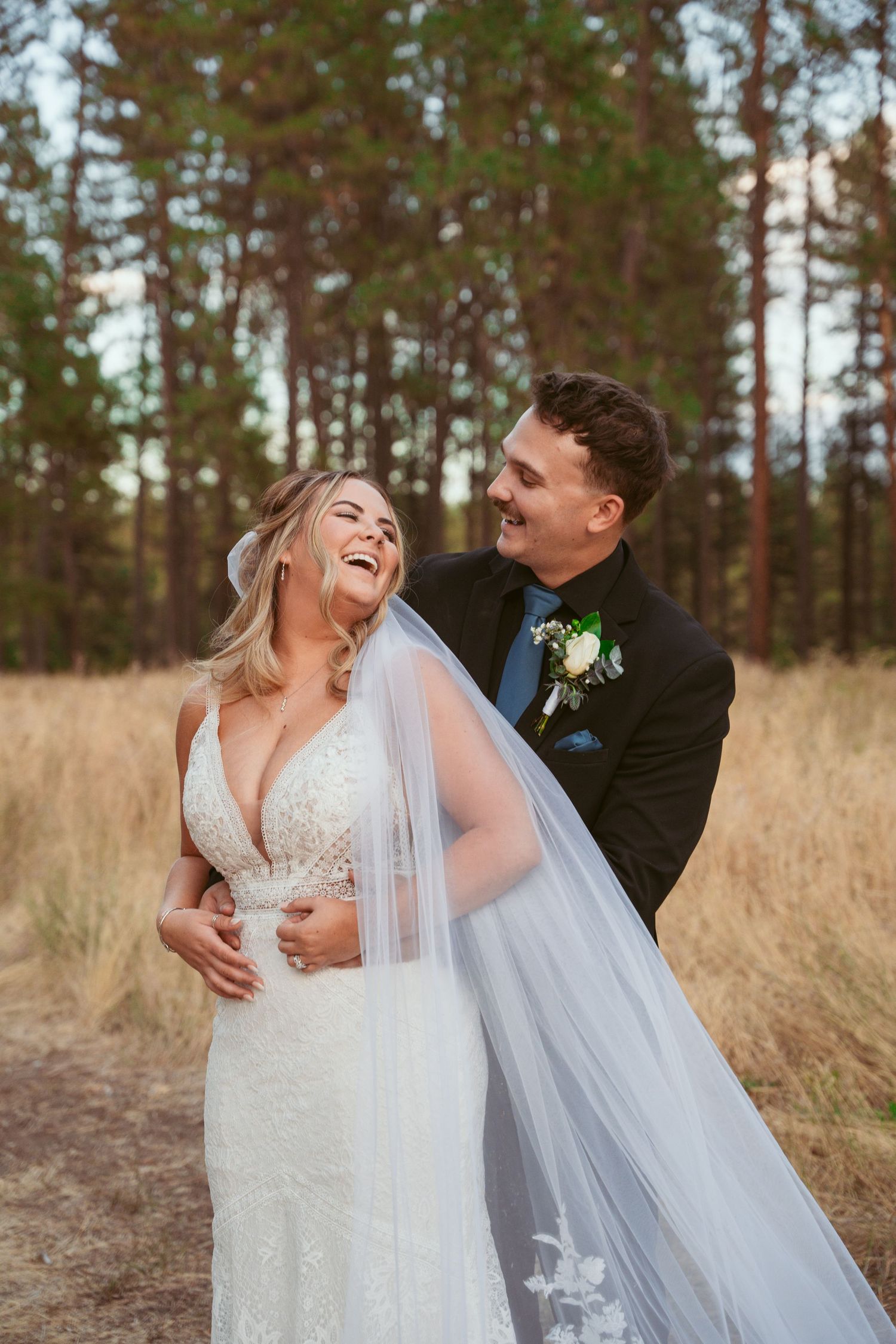 A joyful wedding couple shares a candid moment together in a field with pine trees in the background.