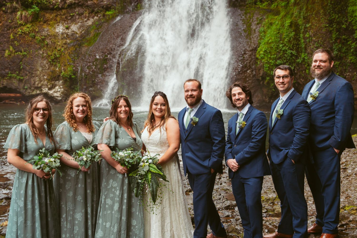 Full wedding party in navy suits and sage dresses pose together by a scenic waterfall.
