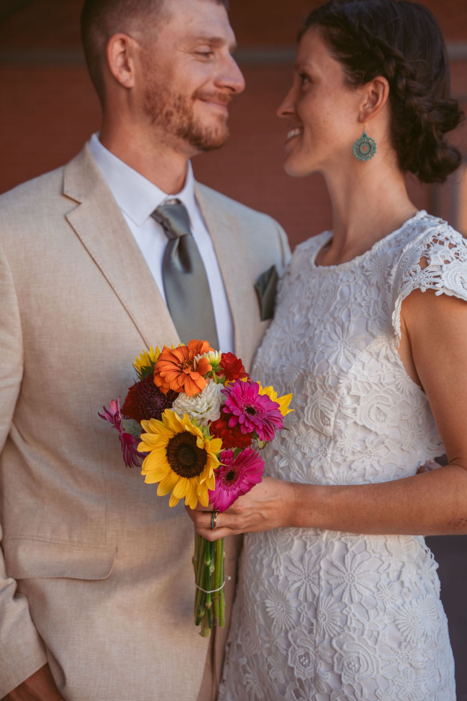 Close-up shots of newlyweds with sunflower bouquet and lace dress details against brick backdrop.