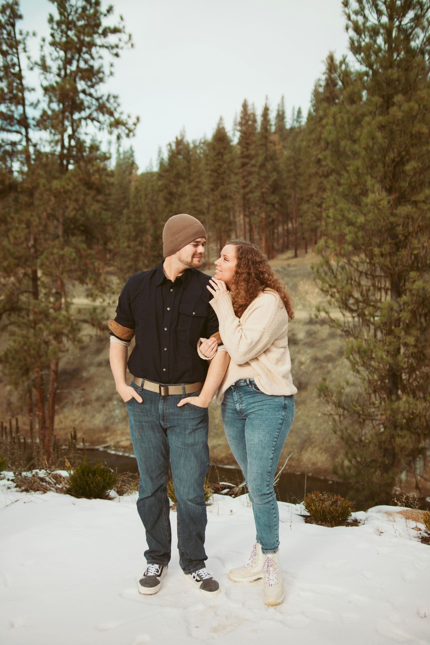 Playful couple laughing together in the snow with pine trees in background.