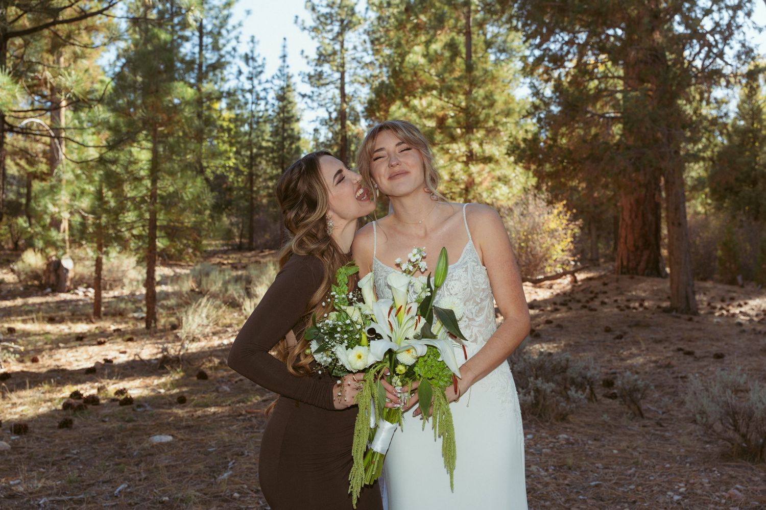 Two people share a joyful moment in a sunlit forest setting while holding white floral bouquets.