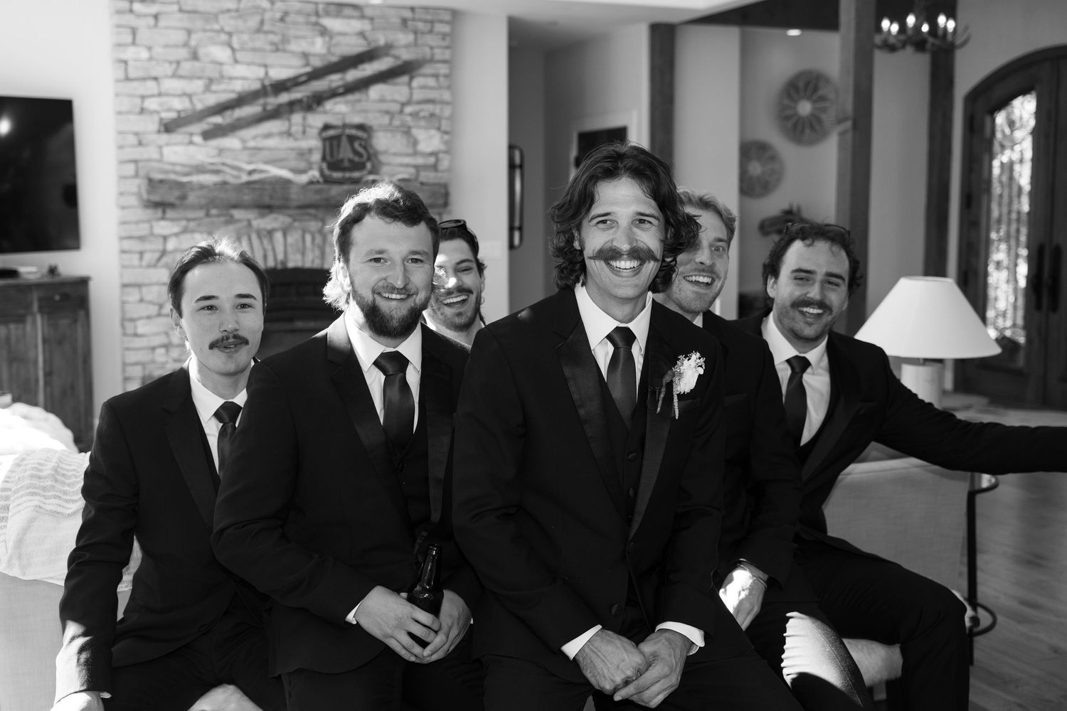 Wedding party members in black suits posing together against a white stone wall.