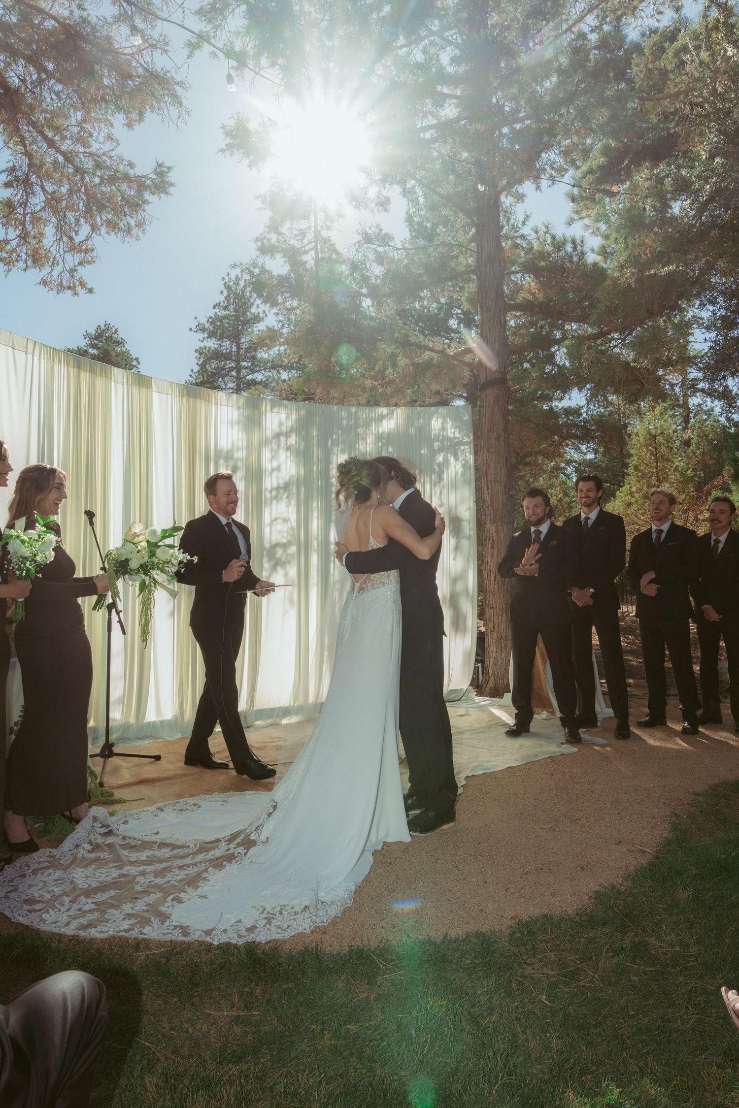 Sun-drenched forest wedding ceremony with white drapes and wedding party in attendance.