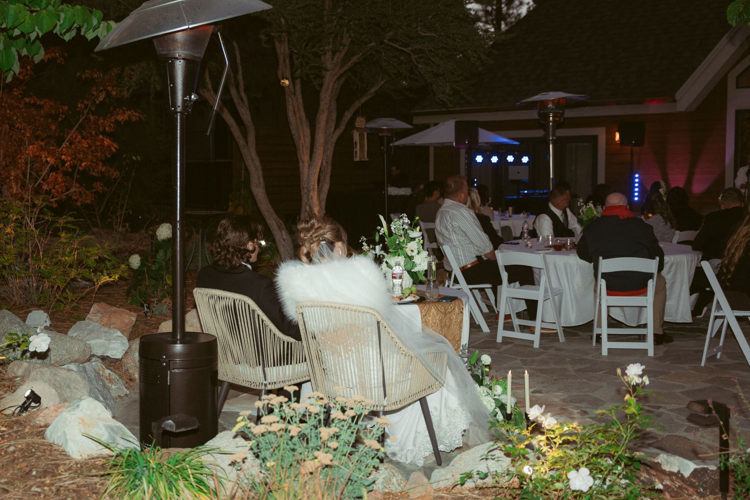 Outdoor wedding reception at night with string lights and white chairs arranged under trees.