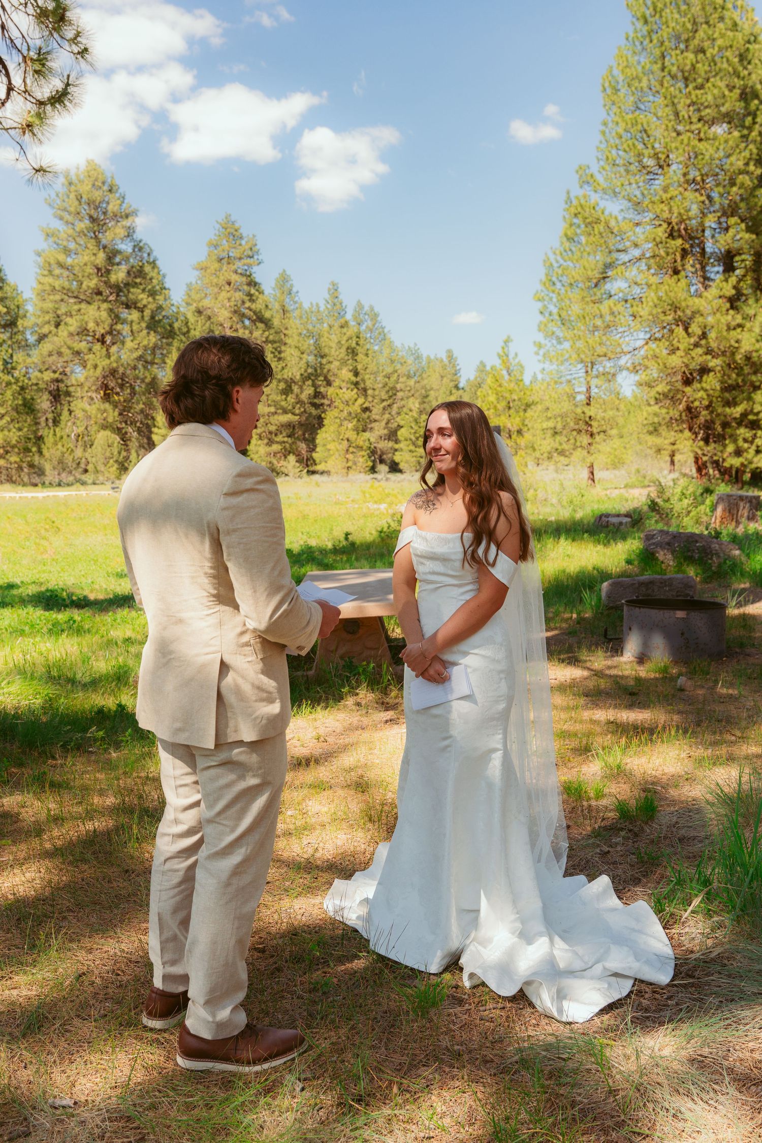 A couple reads personal vows in a sunny mountain meadow surrounded by pine trees.