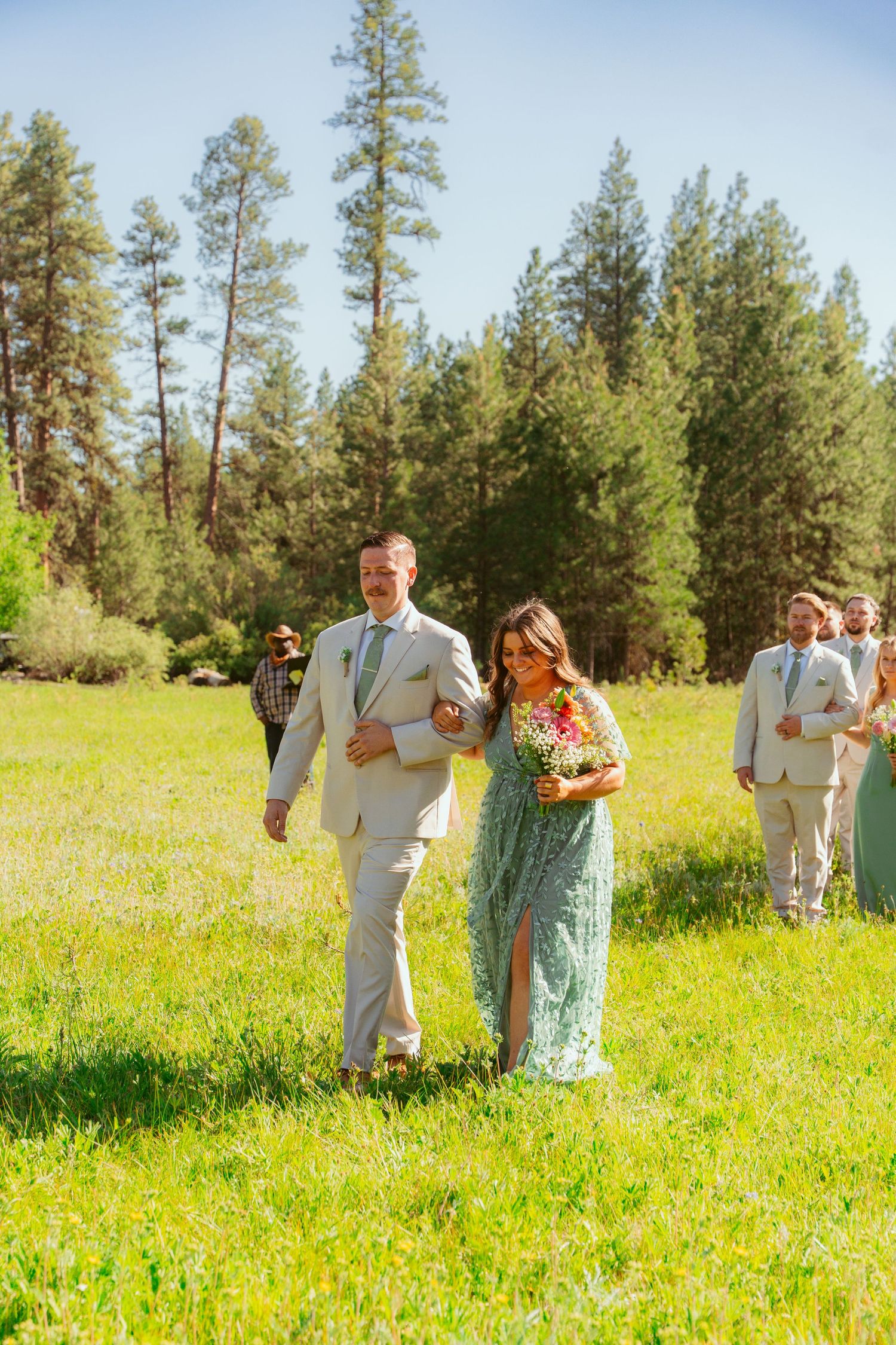 Wedding party members walking through a bright spring meadow with evergreen forest background.