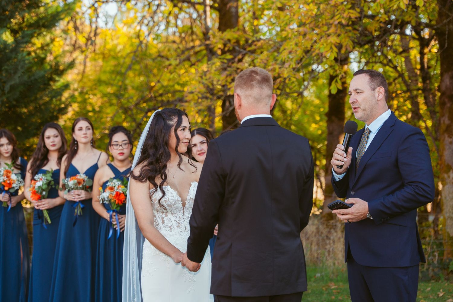 Outdoor wedding ceremony with bridesmaids in navy dresses against golden fall foliage.