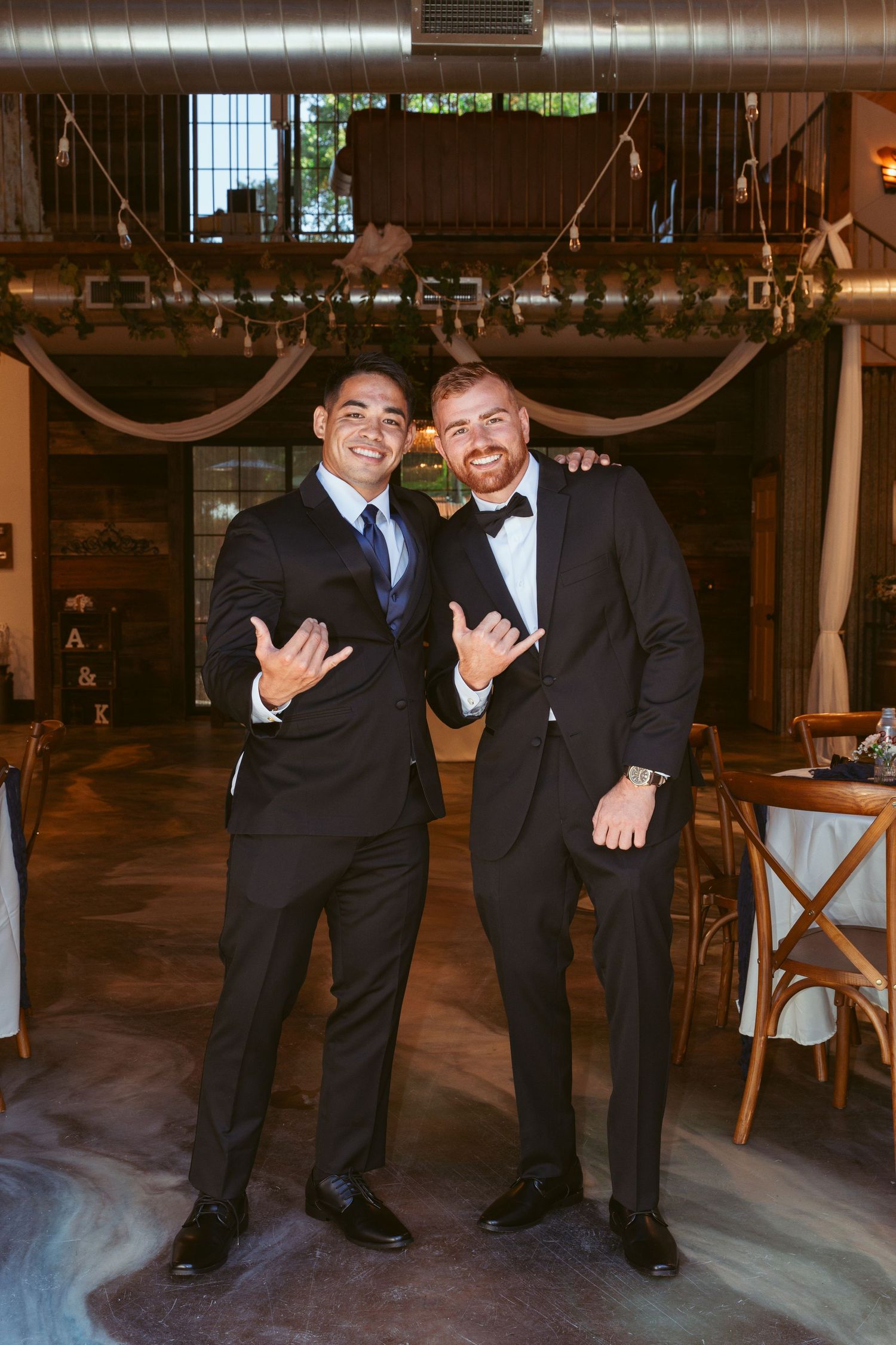 Two wedding guests in formal suits making hand gestures at an indoor reception.