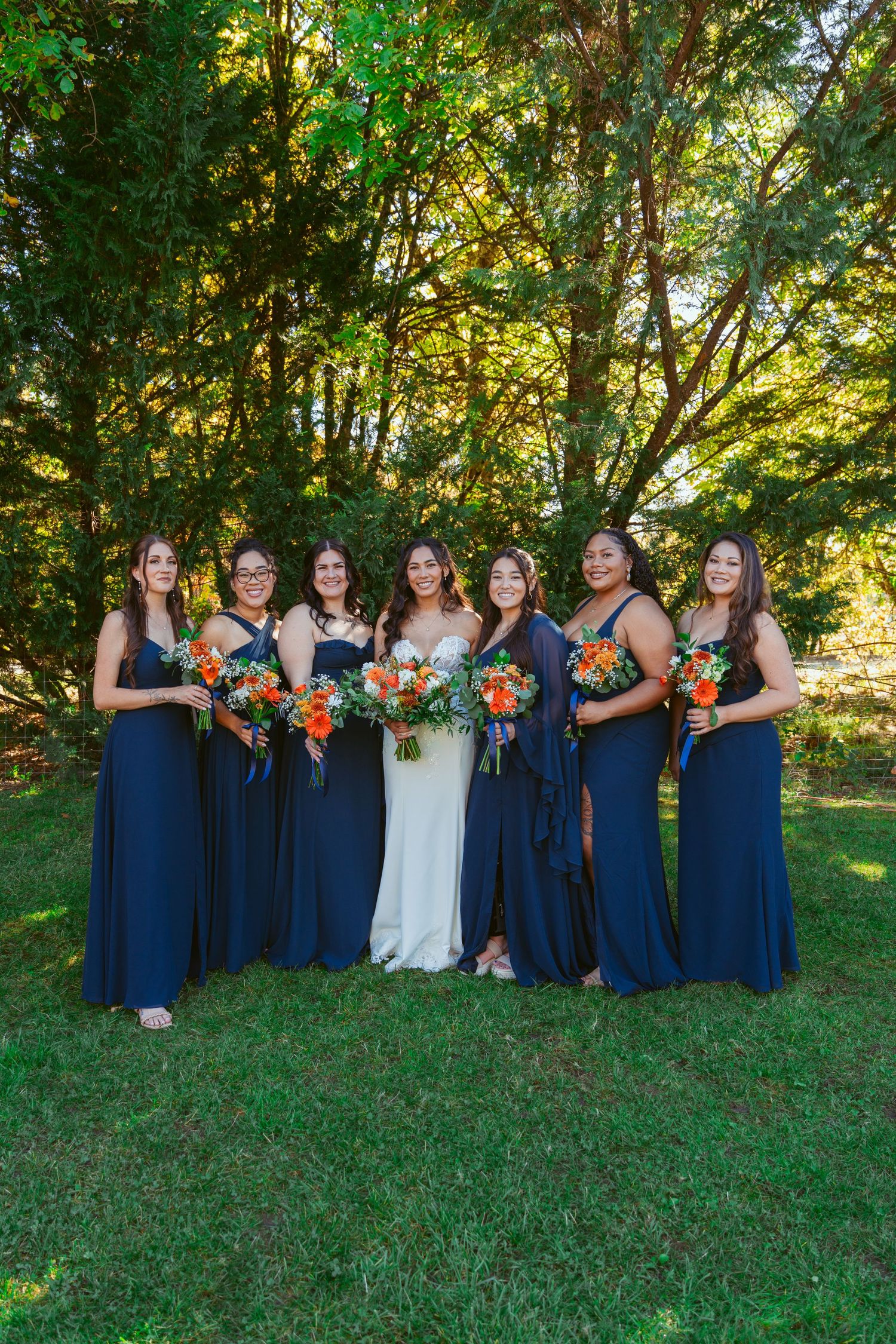 Bridal party in matching navy gowns poses with colorful bouquets against lush green forest backdrop.