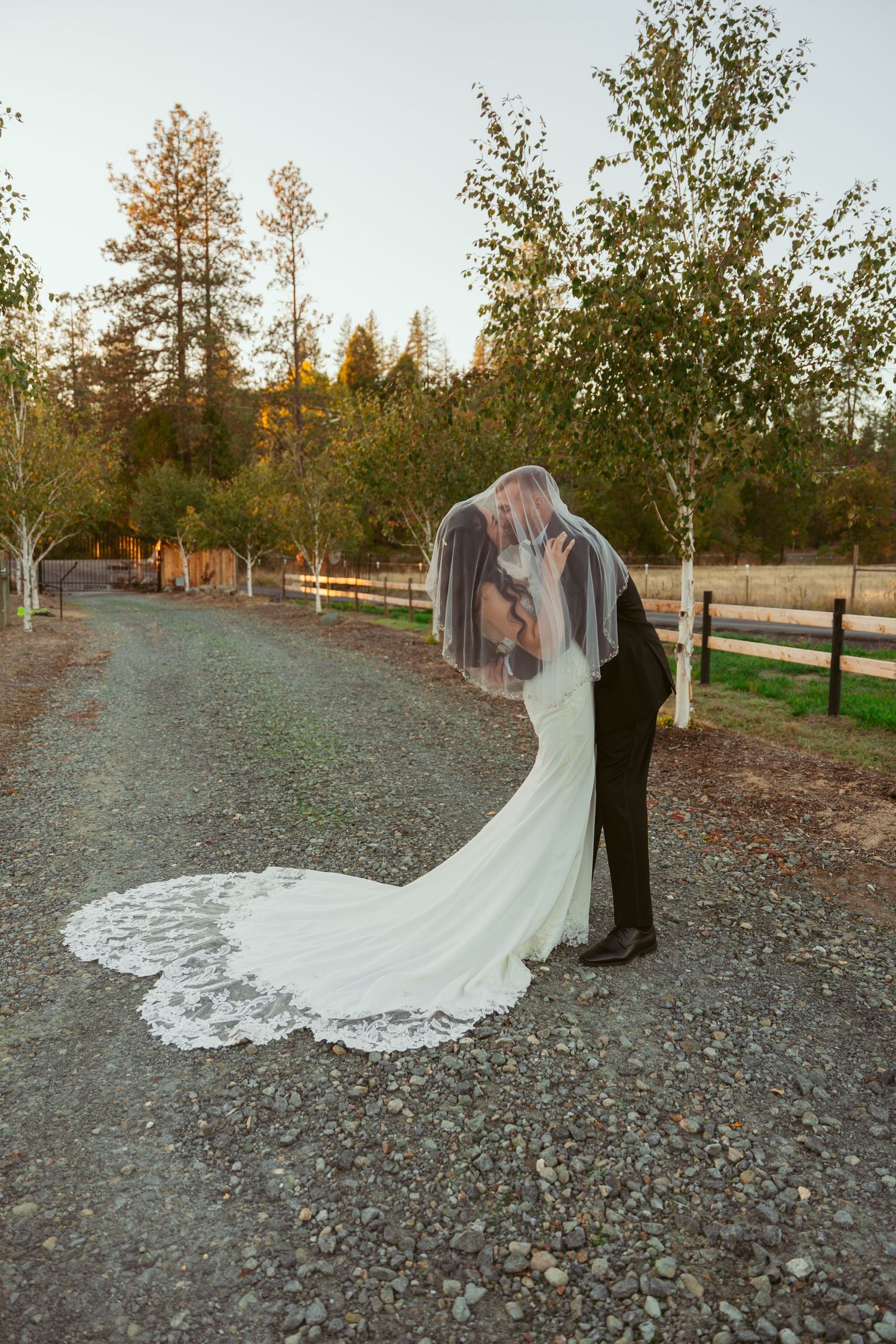 Sunset wedding portrait with dramatic veil and flowing train on gravel path.