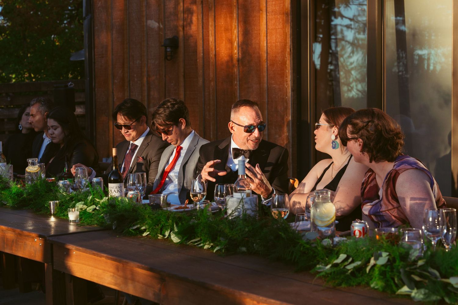 Wedding guests enjoy dinner at rustic wooden tables decorated with greenery under warm lighting.