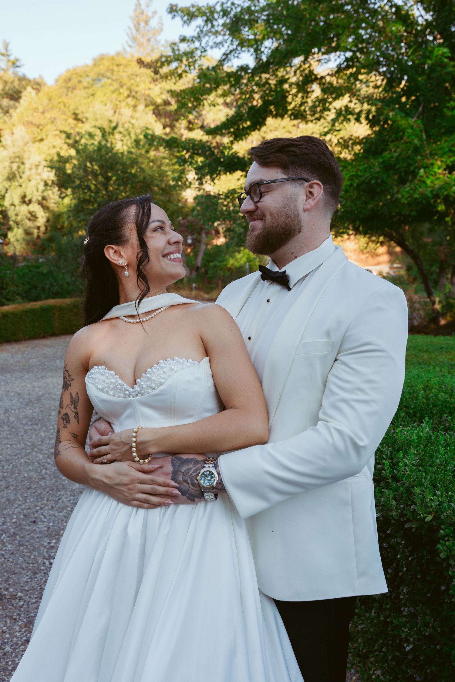 A romantic wedding moment captured outdoors with a couple in white formal attire embracing on a garden path.