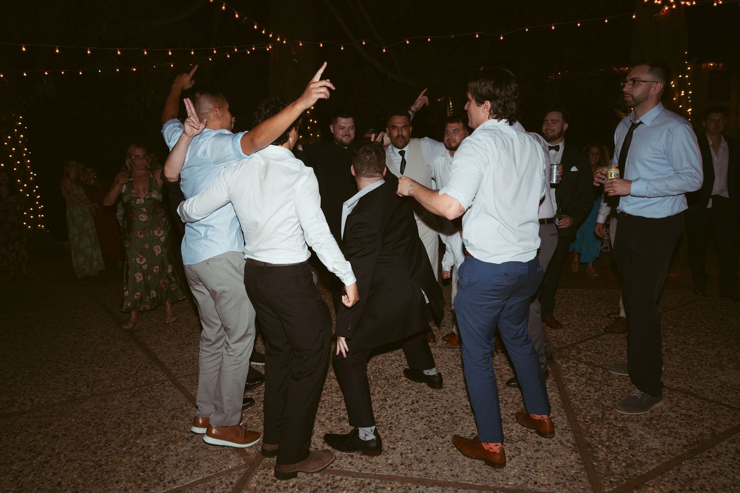 Wedding guests dance together on a dark dance floor during an evening reception celebration.