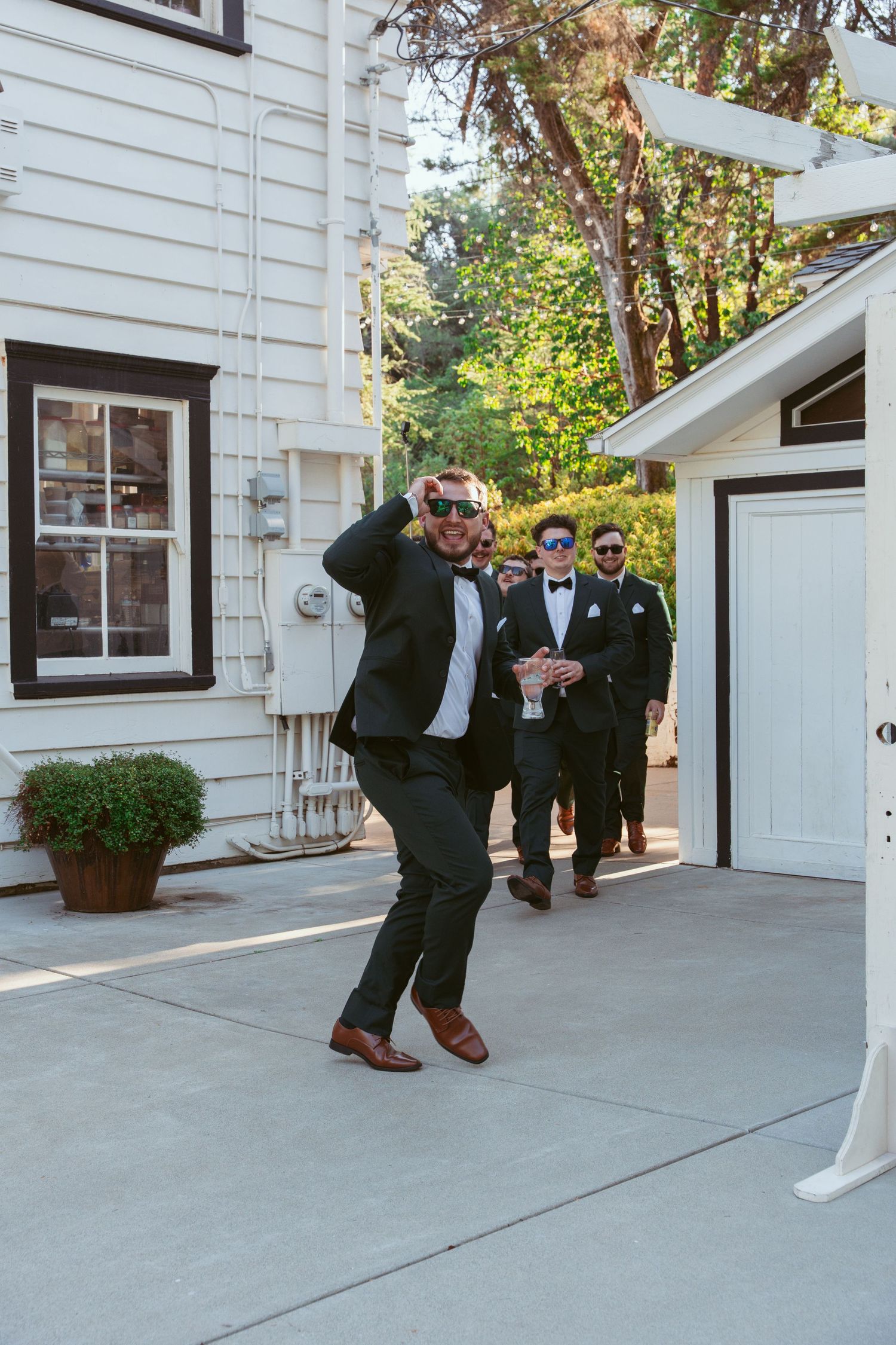 Groomsmen in black tuxedos pose playfully on a concrete patio outside a white building.
