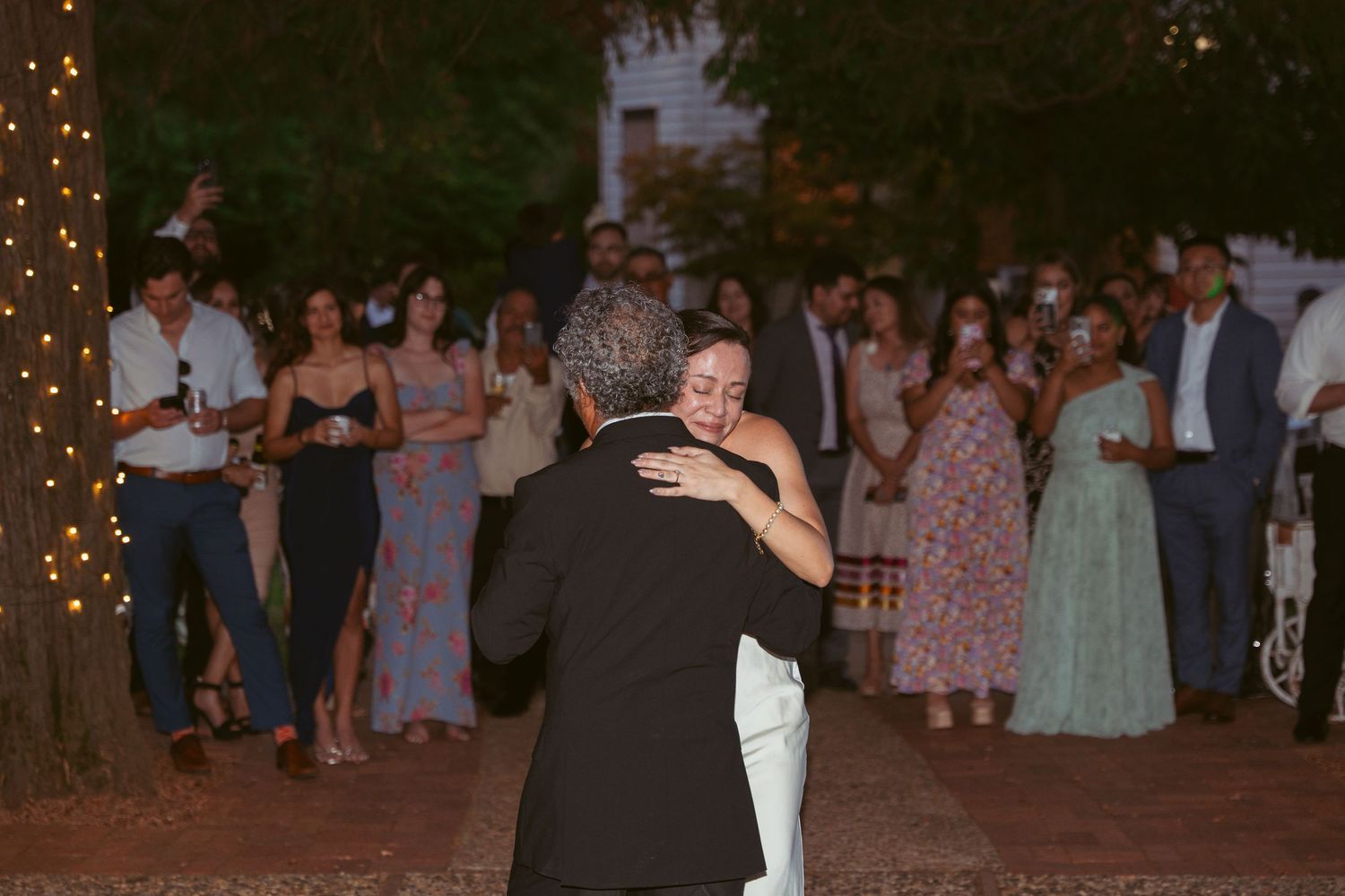 Wedding guests enjoy dancing at a twilight outdoor reception with string lights and trees.