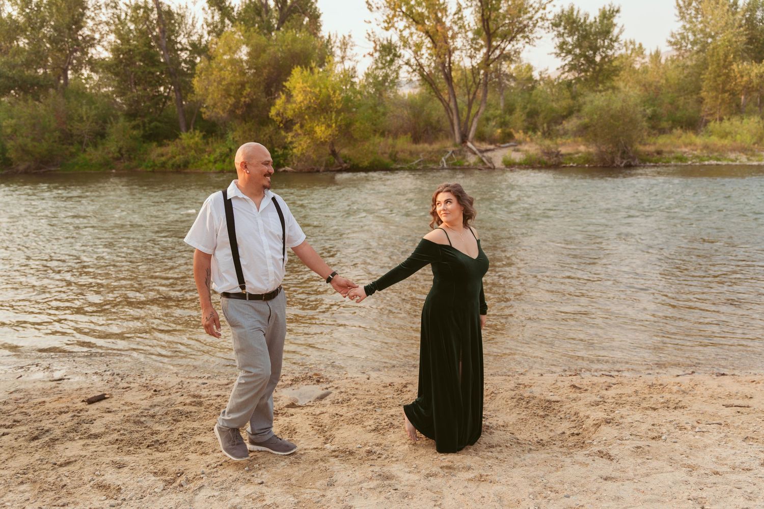 A couple poses together on a sandy beach with water and trees in the background.