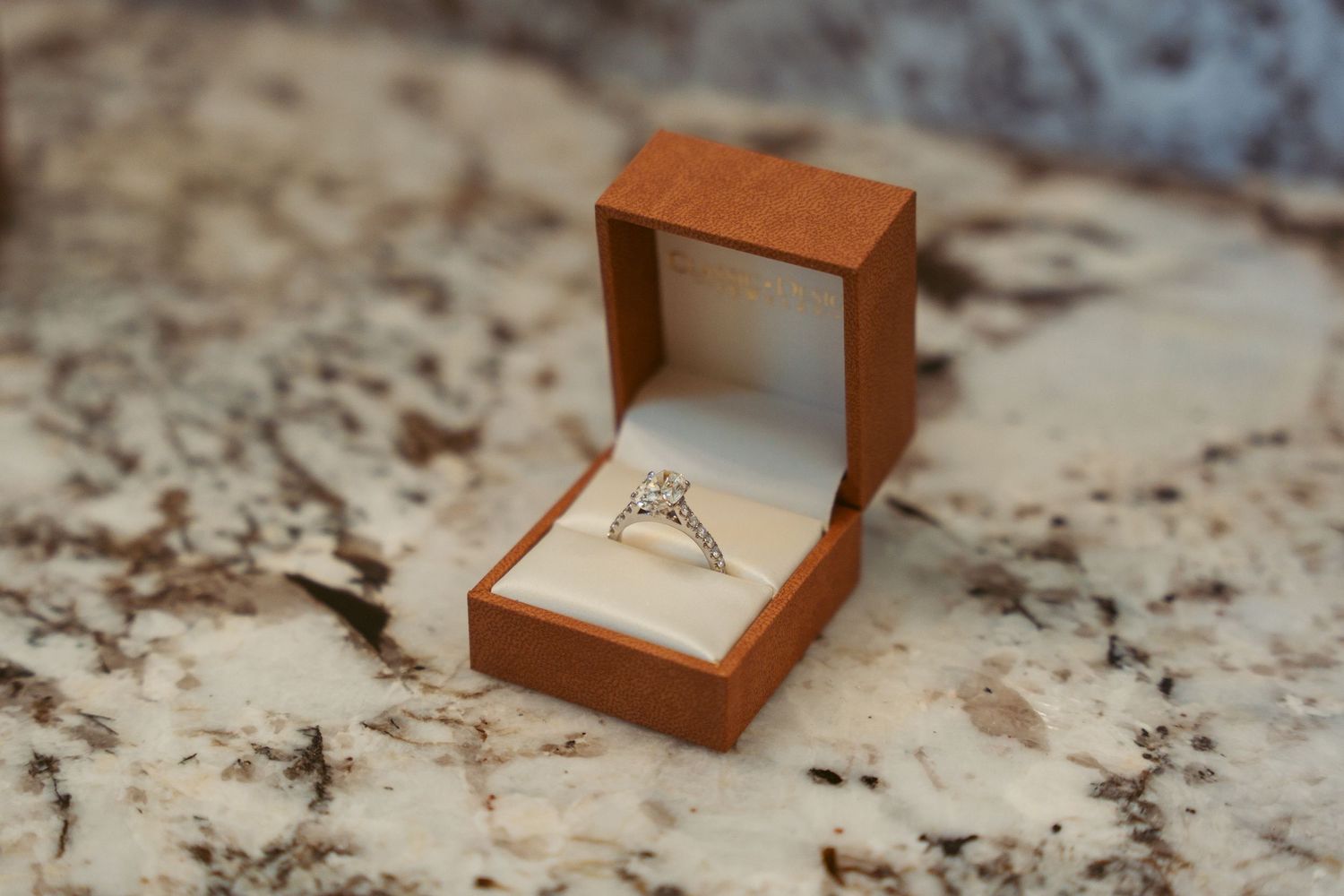 Close-up of an engagement ring in a brown wooden box on a marble countertop.