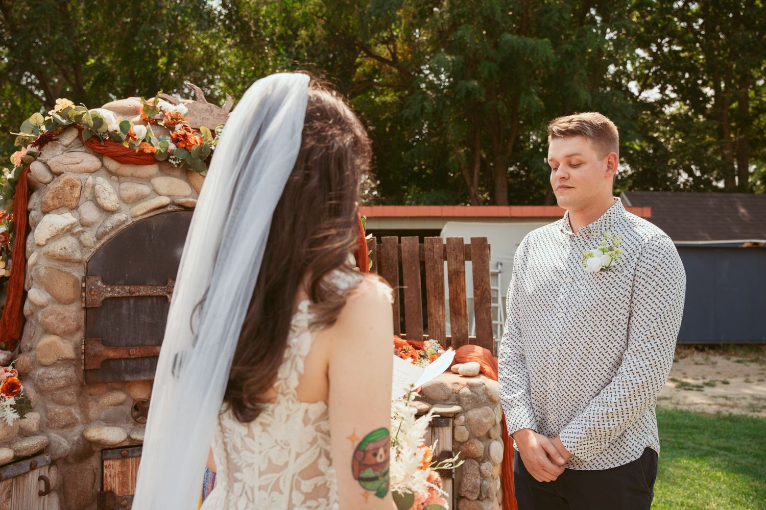 Wedding ceremony participants standing outdoors with a wooden fence and decorative flowers.