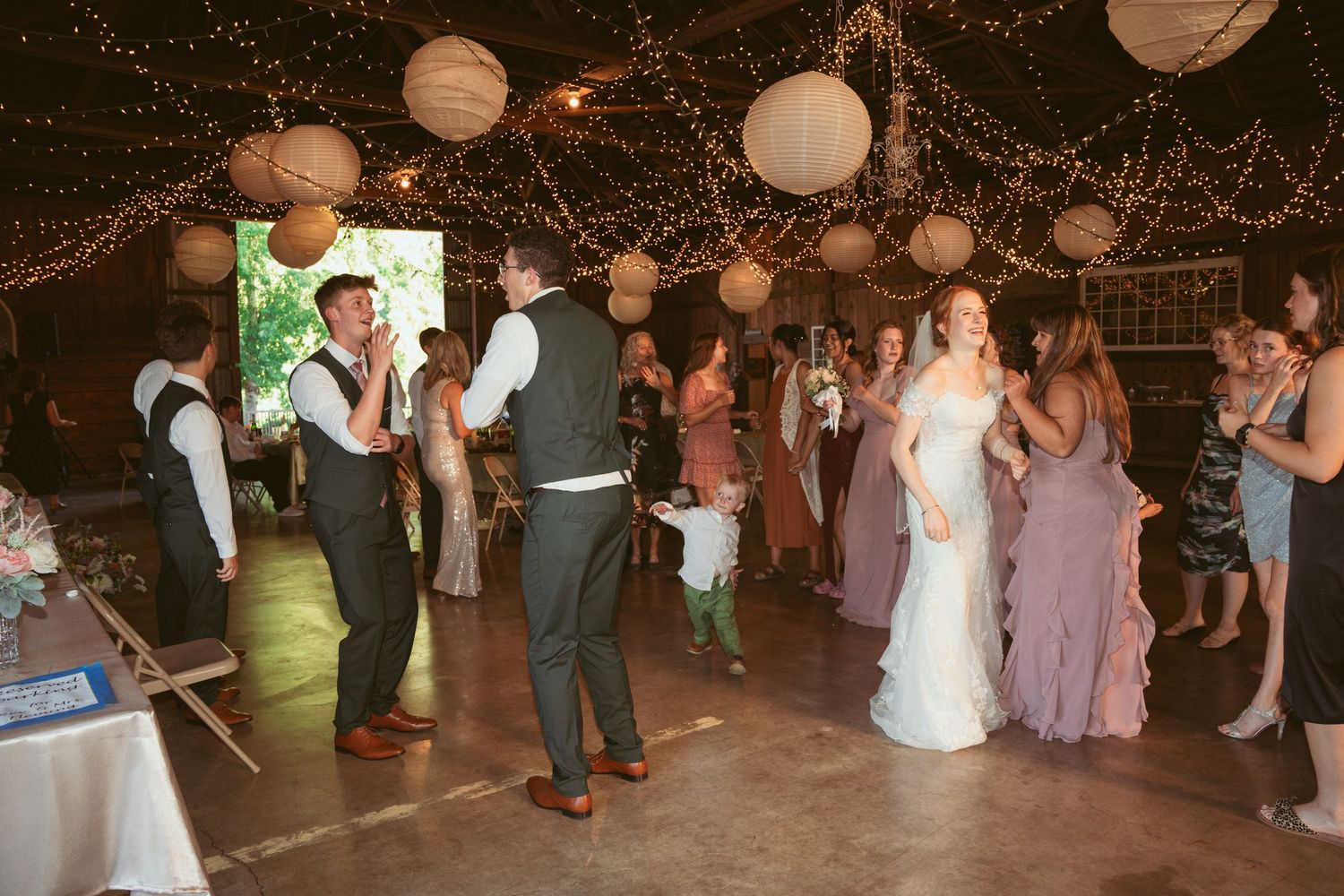 Wedding guests dance under twinkling lights and paper lanterns in a rustic barn reception venue.