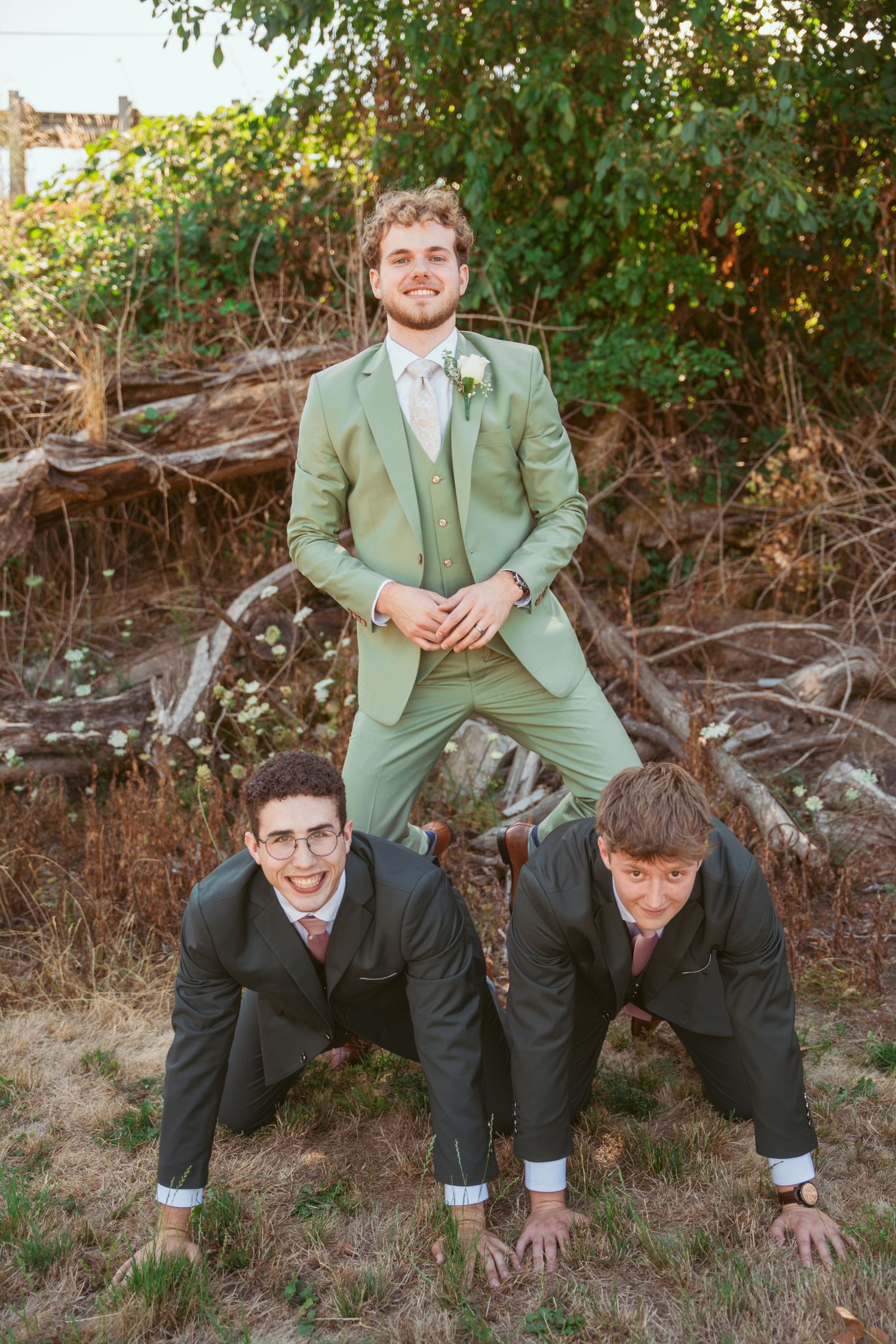 Groomsmen in sage green suits pose casually on outdoor terrain.
