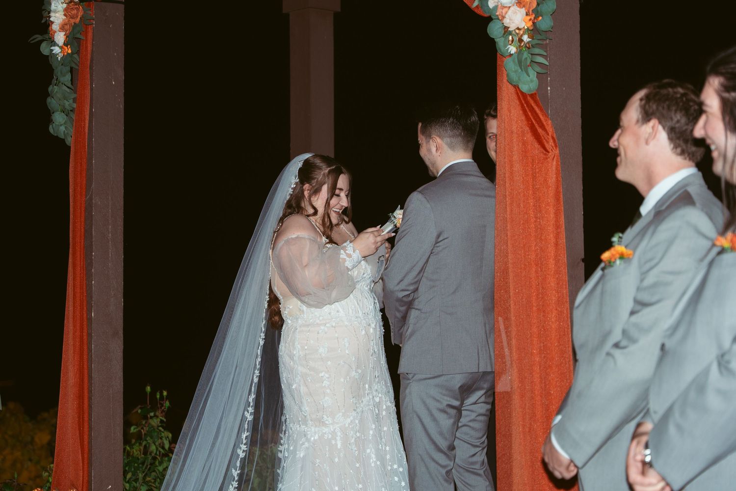 A couple exchanges vows under a red-draped wedding arch with floral decorations at an outdoor evening ceremony.