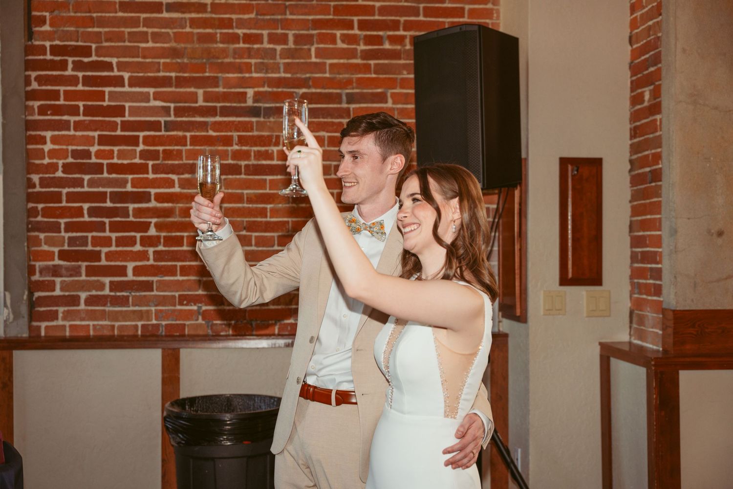 A couple celebrates with champagne glasses at their wedding reception in a rustic brick venue.