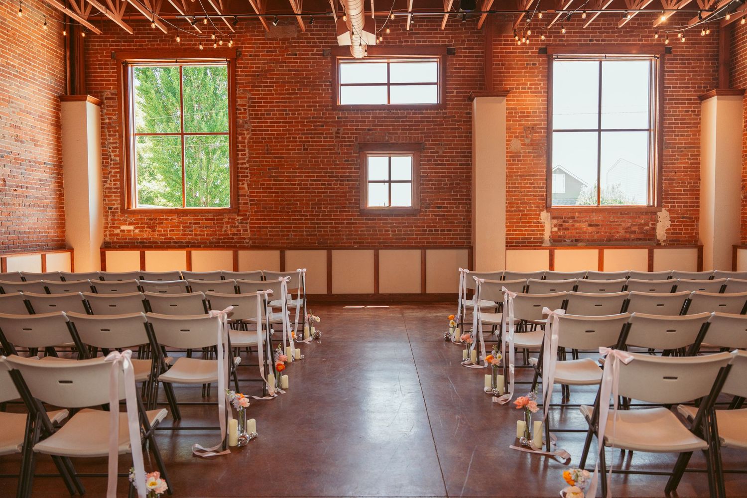 Spacious brick-walled event venue with rows of chairs and string lights overhead.