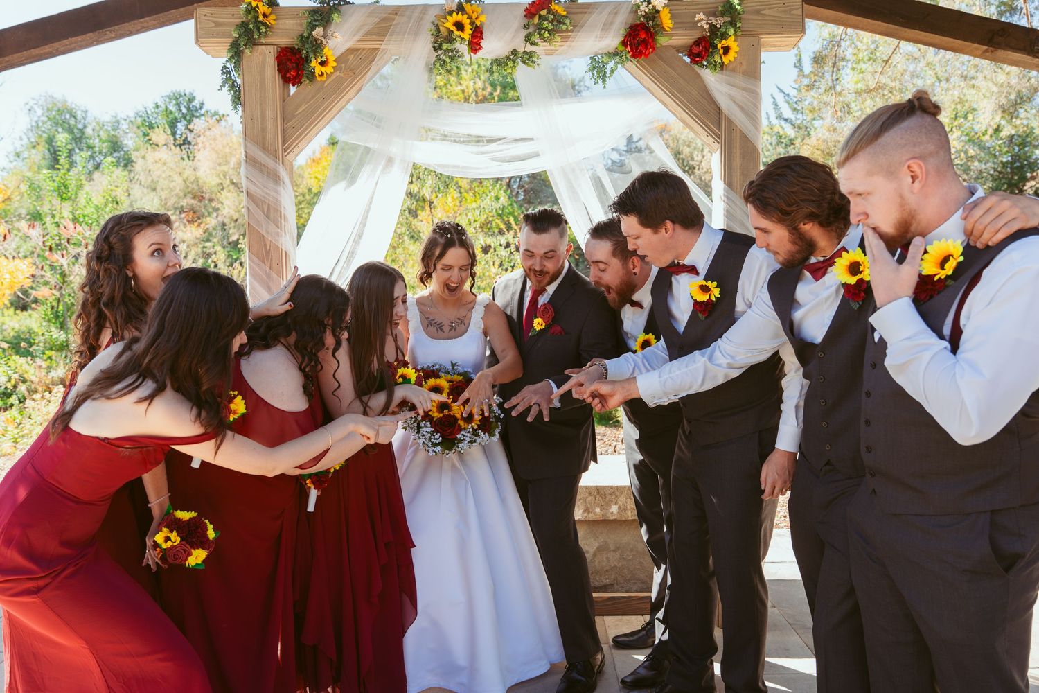 Wedding party in burgundy and black attire poses together under outdoor pavilion.