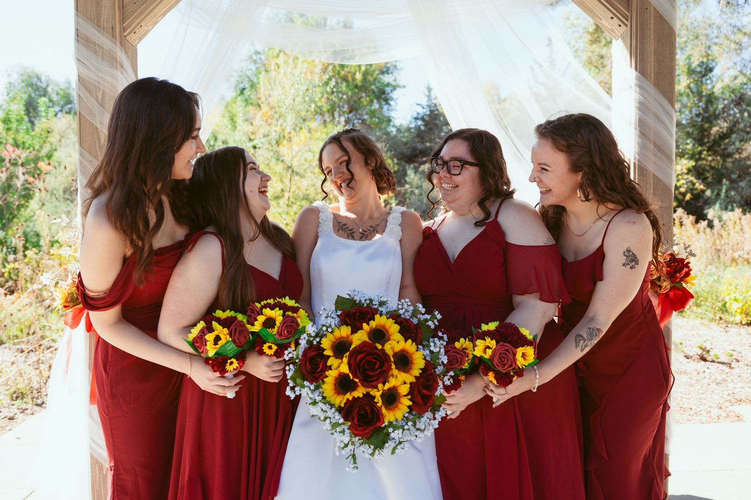 Bridesmaids in burgundy dresses pose with sunflower bouquets under a covered outdoor venue.
