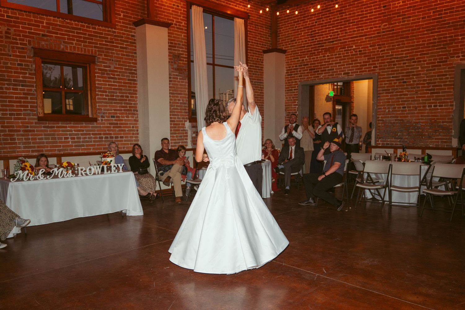 Wedding celebration dance in a rustic brick venue with string lights and white table settings.