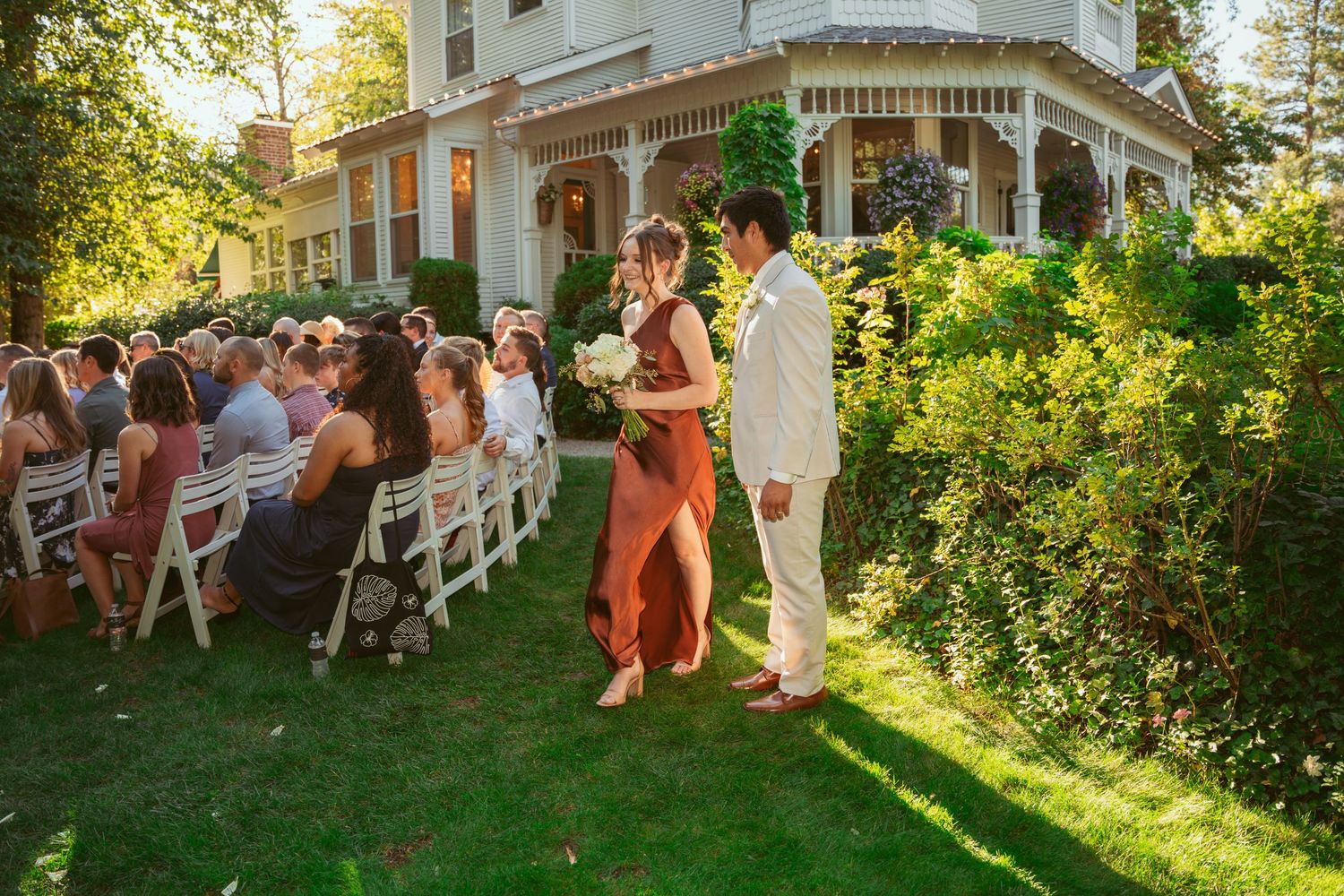 Wedding party and guests mingling on a lawn beside a historic Victorian mansion during golden hour.